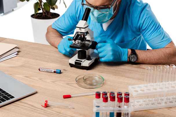 científico en máscara médica y gafas mirando a través del microscopio cerca de tubos de ensayo con letras de coronavirus aislados en blanco
  - Foto, Imagen