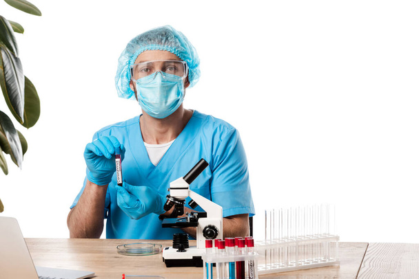 científico en gorra médica y gafas que sostienen el tubo de ensayo con letras de coronavirus cerca del microscopio y el ordenador portátil aislado en blanco
  - Foto, Imagen