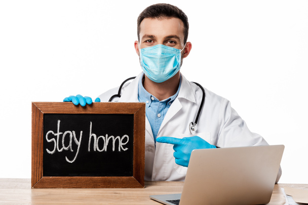 doctor in medical mask and white coat pointing with finger at chalk board with stay home lettering isolated on white  - Фото, изображение