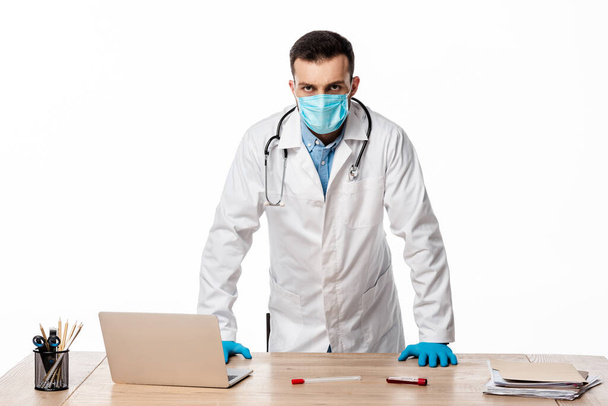 doctor in medical mask and white coat standing near laptop and test tubes isolated on white  - Photo, Image
