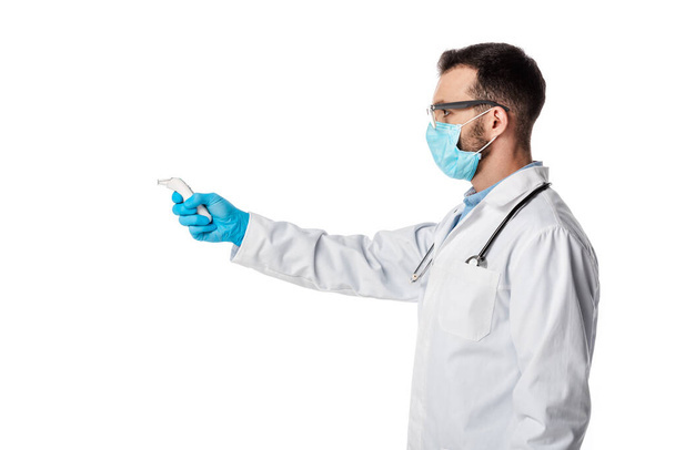 vista lateral del médico en máscara médica y capa blanca sosteniendo pirómetro aislado en blanco
  - Foto, Imagen