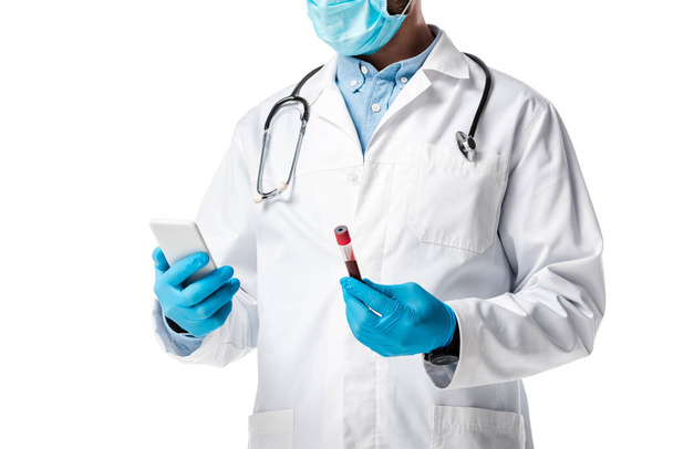 oříznutý pohled na lékaře v lékařské masce a bílém plášti držící smartphone a zkumavku s krevním vzorkem izolovaným na bílém  - Fotografie, Obrázek