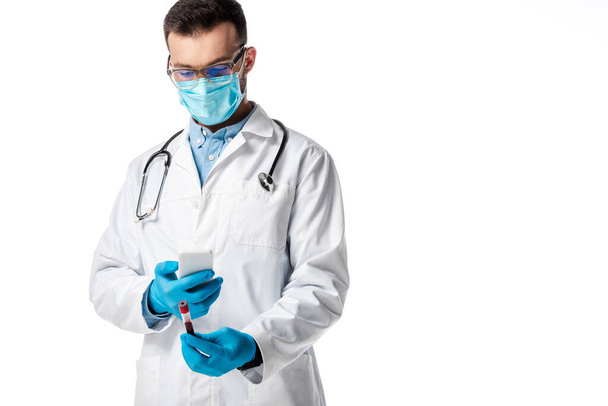 médico con máscara médica y bata blanca que sostiene el teléfono inteligente y toma una foto del tubo de ensayo con muestra de sangre aislada en blanco
  - Foto, Imagen