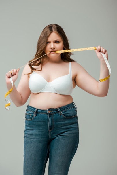 plus size woman in jeans and bra biting measuring tape isolated on grey - Photo, Image