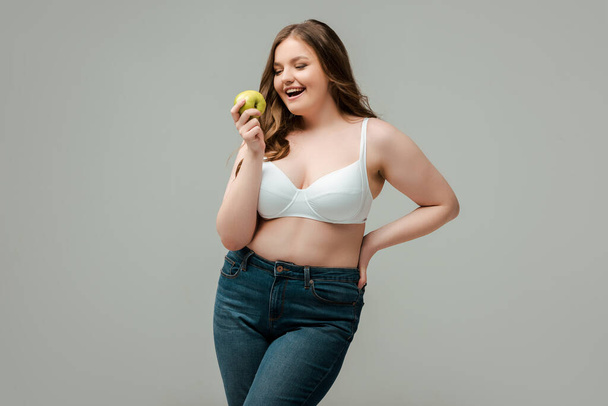 cheerful plus size woman in jeans and bra holding apple isolated on grey - Photo, Image