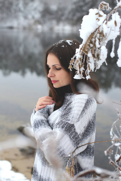 hosszú hajú barna hajú, szürke pulóverben áll egy téli erdő havas partján, kezeit a mellkasára hajtva, fejét lehajtva. - Fotó, kép