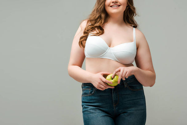 cropped view of cheerful plus size woman in jeans and bra holding apple isolated on grey - Photo, Image