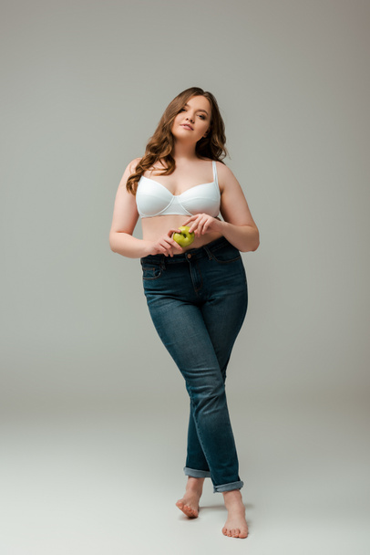 attractive plus size girl in jeans and bra holding apple on grey - 写真・画像