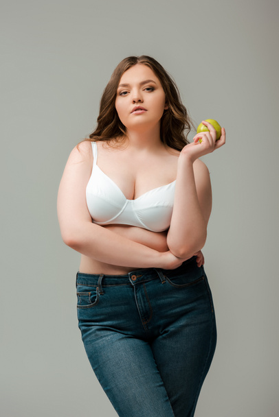 attractive plus size girl in jeans and bra holding apple isolated on grey - Photo, Image