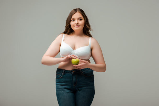 plus size girl in jeans and bra holding apple isolated on grey - Photo, Image