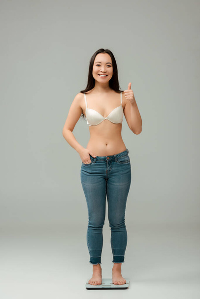 happy overweight asian woman standing on scales and showing thumb up on grey - Fotó, kép