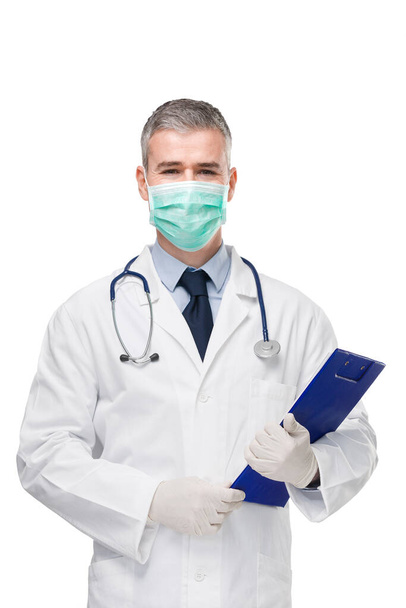 Doctor wearing lab coat, surgical mask, gloves and stethoscope holding a patient file or medical notes on a clipboard in his hands and he  looks at the camera isolated on white - Photo, image