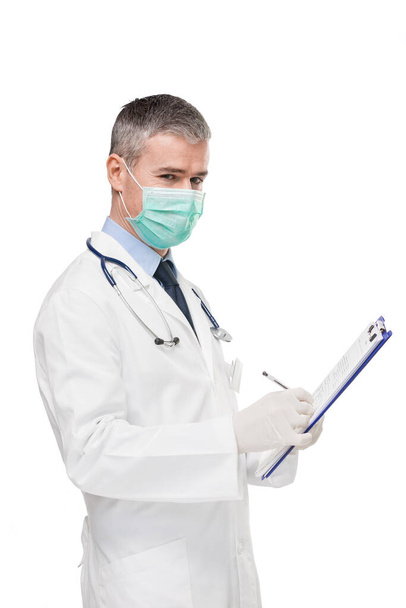 Γιατρός φορώντας εργαστηριακή ποδιά, χειρουργική μάσκα, γάντια και στηθοσκόπιο κρατώντας ένα αρχείο ασθενούς ή ιατρικές σημειώσεις σε ένα πρόχειρο στα χέρια του, όπως ο ίδιος γυρίζει να δούμε την κάμερα που απομονώνονται σε λευκό - Φωτογραφία, εικόνα