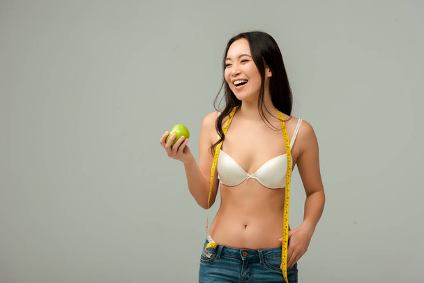 happy and overweight asian girl holding measuring tape and apple isolated on grey  - Photo, Image