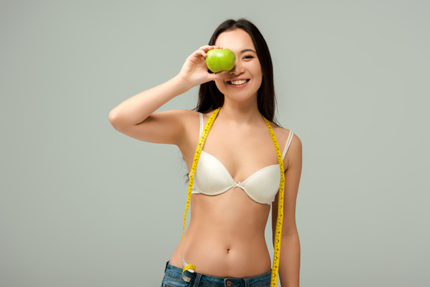 smiling and overweight asian girl holding apple near face isolated on grey  - Photo, Image