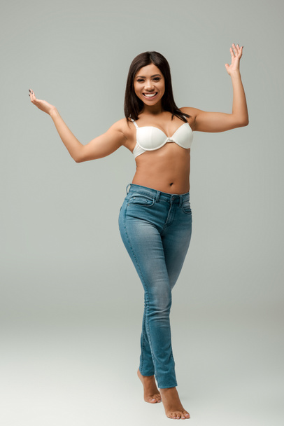 sovrappeso e felice afroamericano ragazza in jeans e reggiseno in piedi su grigio
 - Foto, immagini