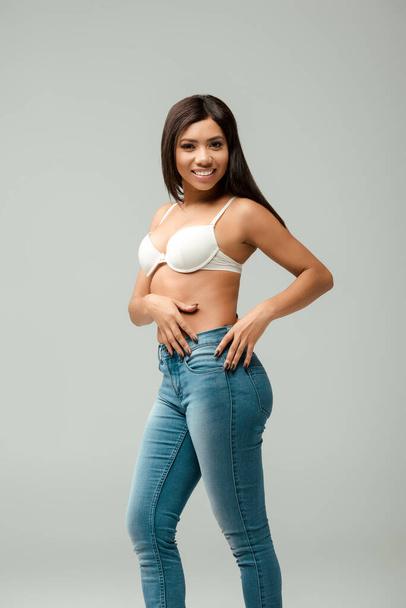overweight and happy african american girl in jeans and bra isolated on grey - Fotó, kép