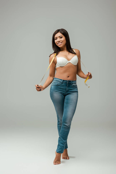 happy and overweight african american girl in jeans and bra holding measuring tape on grey - Foto, Imagem