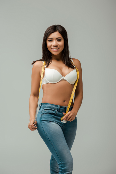 happy and overweight african american girl in jeans and bra holding measuring tape isolated on grey - Fotografie, Obrázek