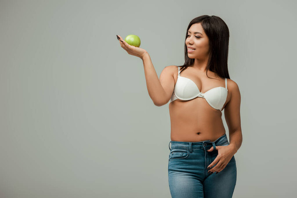 alegre y con sobrepeso afroamericano chica en jeans y sujetador sosteniendo manzana aislado en gris
 - Foto, Imagen