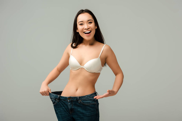 веселая азиатская девушка, указывающая рукой на гигантские джинсы, изолированные на сером
  - Фото, изображение