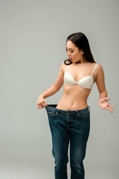 surprised asian woman in oversized jeans isolated on grey  - Foto, Imagem