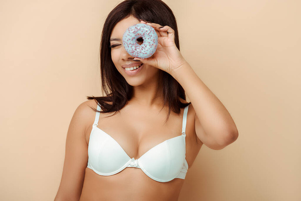 glücklich afrikanisch-amerikanische Mädchen in BH hält Donut in Augennähe isoliert auf beige, Körper positives Konzept  - Foto, Bild