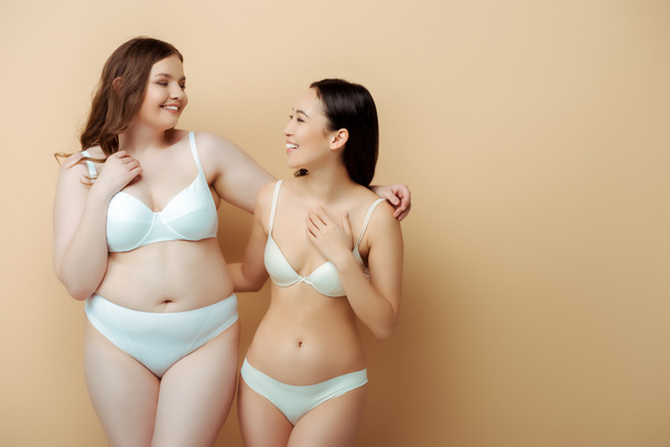 happy plus size woman standing with asian girl in underwear isolated on beige, body positive concept  - 写真・画像