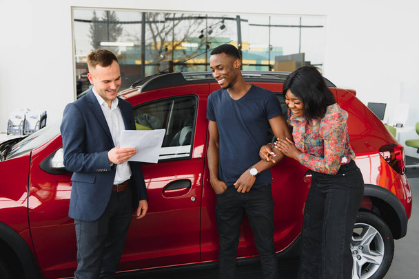 African american family at car dealership. Salesman is showing trunk of new red car. - 写真・画像