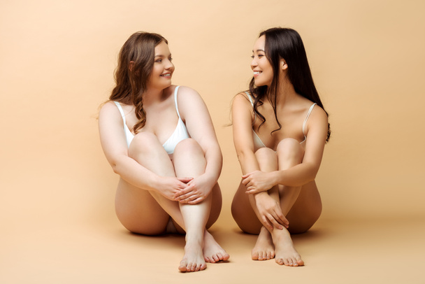 cheerful plus size woman and smiling asian girl in lingerie sitting on beige, body positive concept  - Foto, immagini