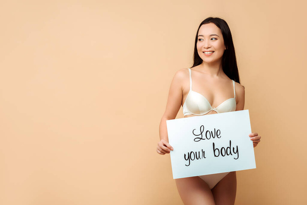 cheerful asian girl holding placard with love your body lettering and looking away isolated on beige  - Foto, Imagem