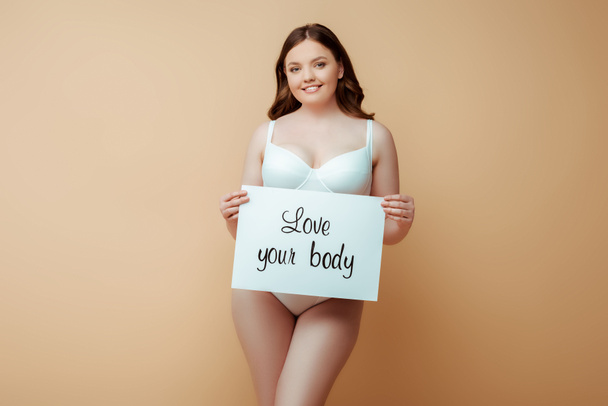 happy plus size girl holding placard with love your body lettering isolated on beige, body positive concept  - Fotó, kép
