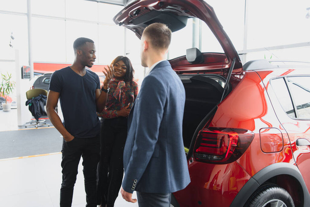African american family at car dealership. Salesman is showing trunk of new red car. - Фото, зображення