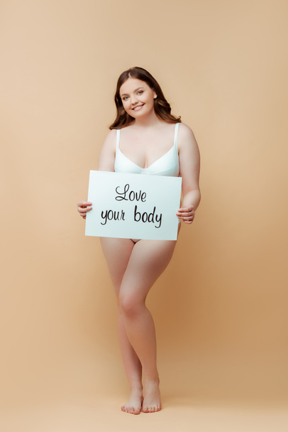 Plus size woman smiling, looking at camera and holding placard with love your body inscription on beige - Photo, Image