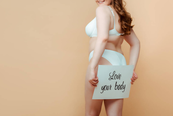 Cropped view of plus size woman presenting placard with love your body inscription isolated on beige - Photo, image