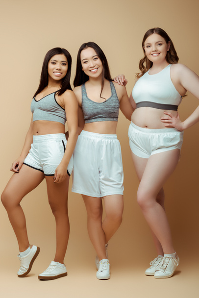 Multiethnic sportswomen smiling and looking at camera on beige - Photo, Image