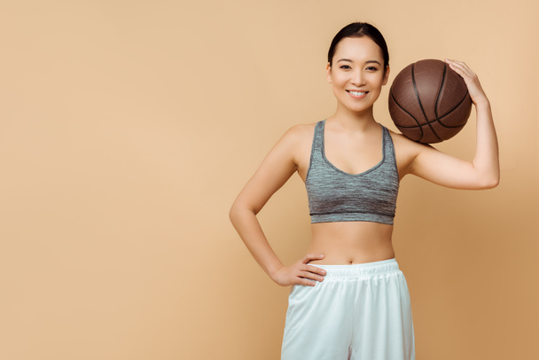 Vista frontale di asiatica sportiva con mano sul fianco e palla sorridente e guardando la fotocamera sul beige
 - Foto, immagini