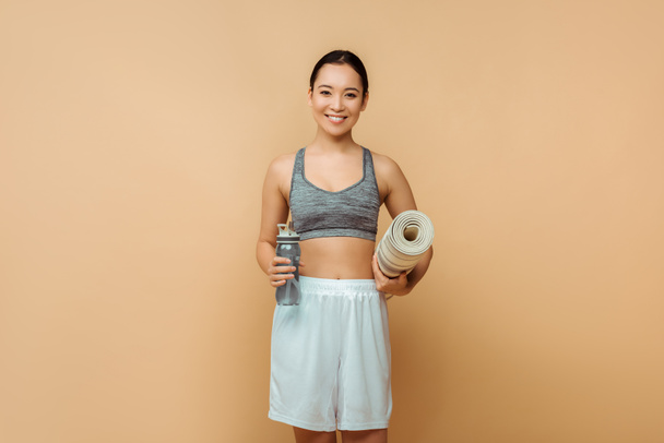 Přední pohled asijské sportovkyně se usmívá, dívá se na kameru a drží sportovní láhev a fitness podložku na béžové - Fotografie, Obrázek