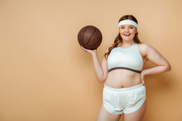 Femme sportive de taille plus avec la main sur la hanche et la balle souriant et regardant la caméra sur beige
 - Photo, image