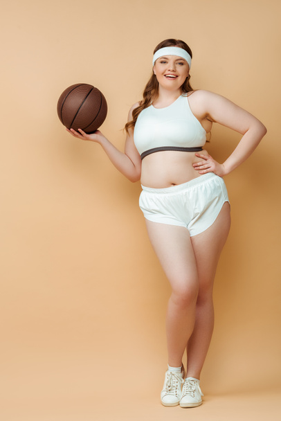 Femme sportive de taille plus avec la main sur la hanche souriant, tenant la balle et regardant la caméra sur fond beige
 - Photo, image
