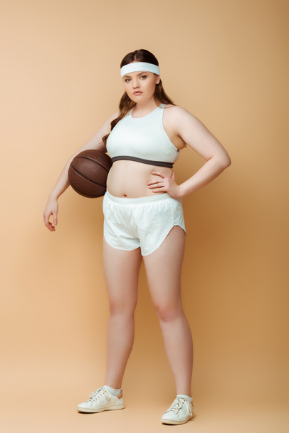 Plus size sportswoman with hand on hip and ball looking at camera on beige - Fotografie, Obrázek