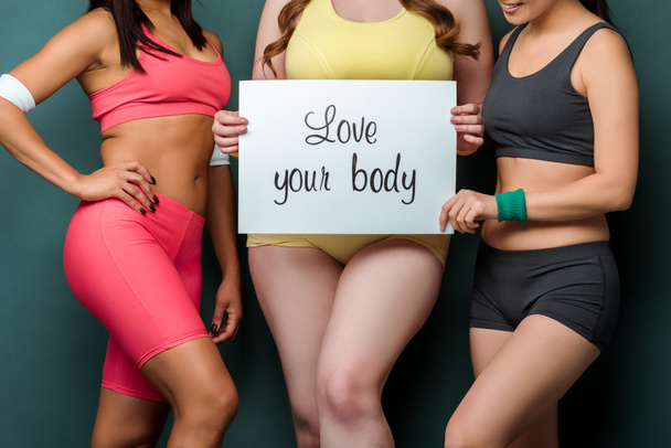 Обрезанный вид мультикультурных спортсменок показывая плакат с любовью ваше тело буквы на зеленом фоне
 - Фото, изображение