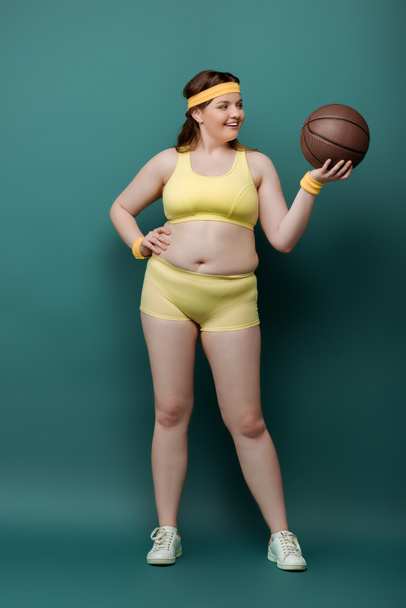Plus size sportswoman with hand on hip looking at ball and smiling on green background - Photo, Image