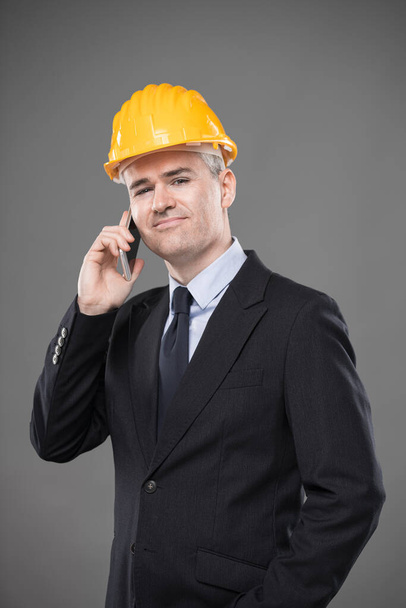 Дружелюбный улыбающийся архитектор или инженер-строитель в шикарном костюме, галстуке и каске, стоящий в чате на мобильном телефоне и смотрящий в камеру с сияющей улыбкой, изолированной на сером
 - Фото, изображение