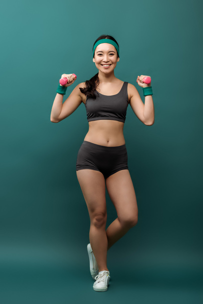 Happy asian sportswoman training with dumbbells smiling and looking at camera on green background - Foto, Bild