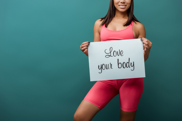 Cropped view of african american sportswoman smiling and holding placard with love your body inscription isolated on green - Foto, Bild