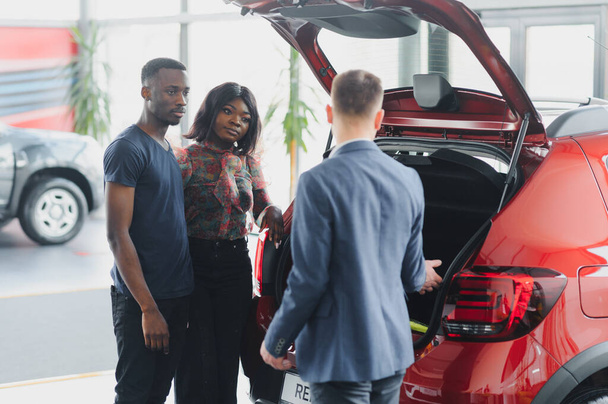 African american family at car dealership. Salesman is showing trunk of new red car. - Foto, immagini