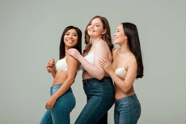 Glückliche multikulturelle Frauen lächeln und umarmen isoliert auf grau - Foto, Bild