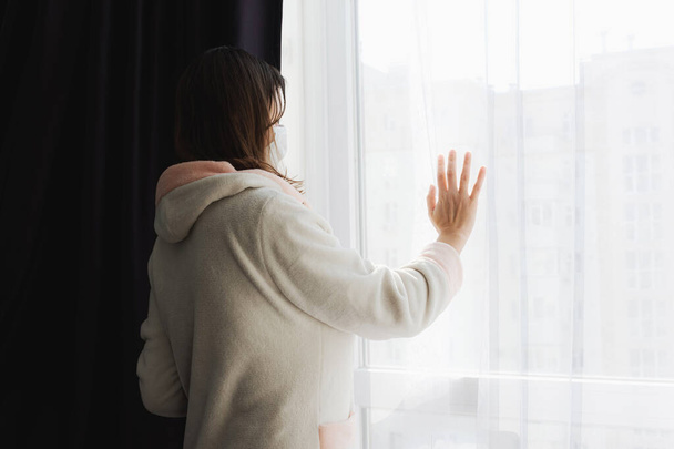 Een verdrietig meisje kijkt uit het raam en raakt het glas aan met haar hand. Een meisje tijdens de quarantaine als gevolg van de coronavirus pandemie kijkt uit door een raam. Isolatie en zelfisolatie. - Foto, afbeelding