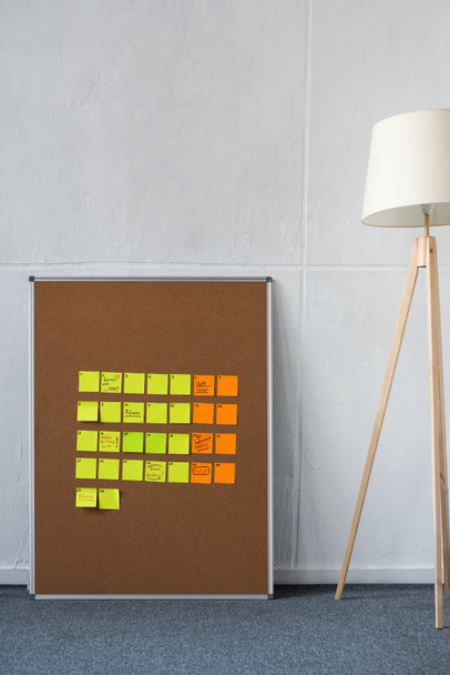 Bruin bord met gele en oranje plakkerige notities met vloerlamp bij muur - Foto, afbeelding